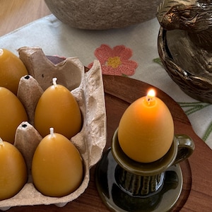 May include: An egg-shaped candle is lit, sitting on a dark green teacup-shaped holder. Several unlit egg-shaped candles are in a cardboard egg carton. The candles are a warm yellow color, and the scene is set on a wooden tray.
