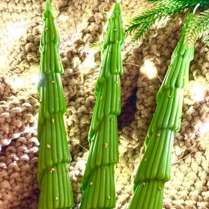 Puede incluir: Tres velas verdes con forma de árbol de Navidad con detalles dorados. Las velas están dispuestas sobre una superficie beige texturizada, con algunas ramitas de pino y luces de hadas. Las velas tienen un diseño escalonado.