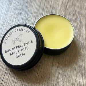 Puede incluir: Lata negra abierta de Mixology Candle Co. Bug Repellent & After-Bite Balm. El bálsamo es de color amarillo claro. La lata está abierta, revelando el bálsamo en su interior. La lata está sobre una superficie de madera.