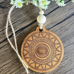 Puede incluir: Un adorno de madera con un diseño de mandala marrón, colgado de una cuerda de yute con una cuenta de madera blanca.