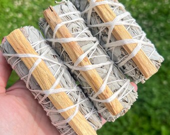May include: Three white sage smudge sticks bundled together with palo santo wood. The sticks are tied with white string and are ready to be used for smudging.