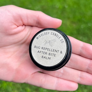 Puede incluir: Una pequeña lata redonda negra de "Bug Repellent & After-Bite Balm" de Mixology Candle Co. se sostiene en una mano. La etiqueta es blanquecina con texto negro y una ilustración de mosquito. El fondo es hierba verde borrosa.