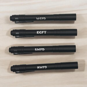 Puede incluir: Cuatro bolígrafos negros con letras plateadas. Los bolígrafos están etiquetados con WCFD, ECFT, SWFD y NWFD.