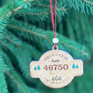 May include: A wooden Christmas ornament with the text "There's no place like home for the holidays" and the zip code 46750. The ornament is round with a light brown finish, blue trees, and a red ribbon for hanging.