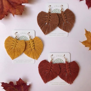 May include: Three pairs of handmade macrame earrings in brown, yellow, and red. Each pair is shaped like a heart with fringe details. The earrings are displayed on cards with the text "Eco Friendly Handmade Macrame Jewellery".