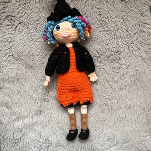 May include: A crocheted witch doll with blue and pink hair, wearing a black hat, black jacket, orange dress, and brown shoes.