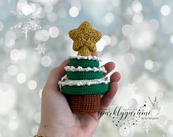 May include: A small crocheted Christmas tree with a gold star on top. The tree is green and white with a brown base. The tree is being held in a hand with a gold bracelet.