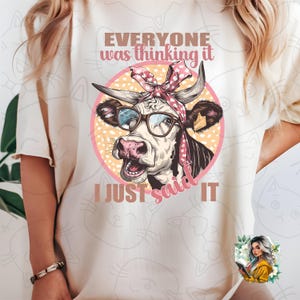 Könnte beinhalten: Weißes T-Shirt mit einer Comic-Kuh, die eine Brille und ein rosa Band trägt. Der Text auf dem Shirt lautet "Everyone was thinking it I just said it."