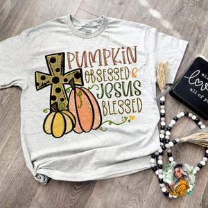 May include: A gray t-shirt with a graphic design featuring a pumpkin, a cross with black polka dots, and the text "Pumpkin Obsessed & Jesus Blessed".