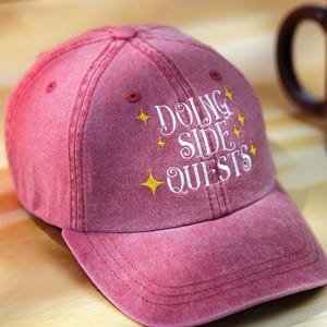 May include: A red, pigment-dyed baseball cap featuring white embroidered text that reads "DOING SIDE QUESTS" surrounded by small, yellow, four-pointed star icons. The cap has a curved brim, a button on top, and stitched eyelets for ventilation.