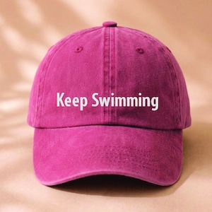 May include: A magenta baseball cap with a structured crown and a curved brim. The front of the cap features the text Keep Swimming printed in white, sans-serif capital and lowercase letters centered across the two front panels of the hat.