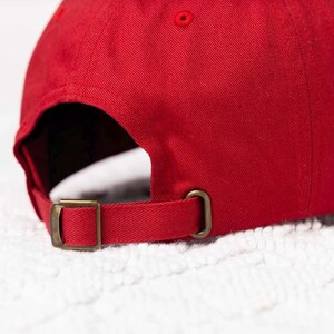May include: A red baseball cap with a brown adjustable strap and buckle. The cap is made of a soft, cotton fabric.