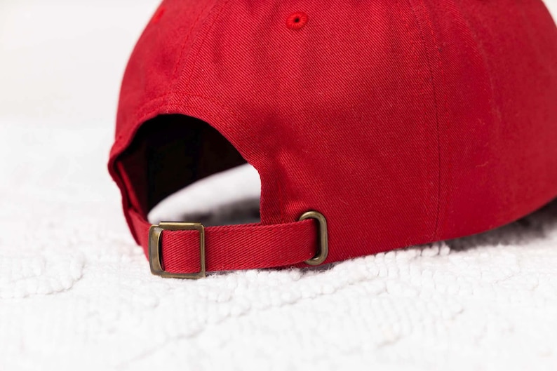 May include: A red baseball cap with a brown adjustable strap and buckle. The cap is made of a soft, cotton fabric.