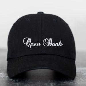 Op de afbeelding: Zwarte baseballcap met de geborduurde tekst "Open Book" in witte letters. De cap heeft een gebogen klep en een knoop aan de bovenkant. Een modieus accessoire voor dagelijks gebruik.