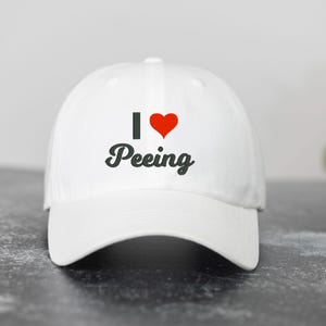 May include: A white baseball cap featuring the text I, a red heart symbol, and the word Peeing in a black cursive font centered on the front panel. The cap has a structured crown, a curved brim, and a button at the top of the dome.