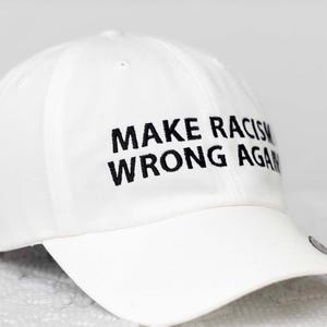 May include: White baseball cap with black embroidered text that reads "Make Racism Wrong Again."