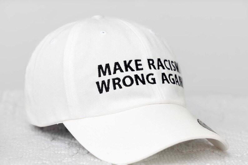 May include: White baseball cap with black embroidered text that reads "Make Racism Wrong Again."