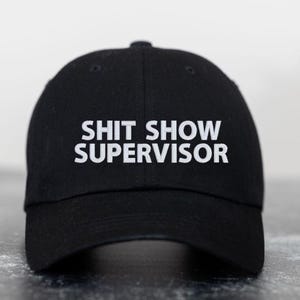 May include: Black baseball cap with white embroidered text that reads "SHIT SHOW SUPERVISOR".