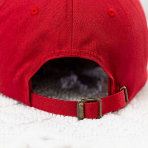 May include: Red baseball cap with a brown adjustable strap and buckle. The cap is made of a soft, cotton fabric.
