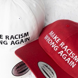 May include: Two baseball caps, one white and one red, both with the text "Make Racism Wrong Again" embroidered in black and white respectively.