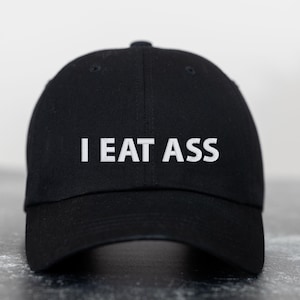 May include: A black baseball cap with a white embroidered phrase "I EAT ASS".