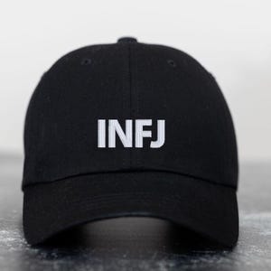 May include: Black baseball cap with the embroidered letters "INFJ" in white. The cap has a curved brim and a button on top. The hat is made of a soft fabric.