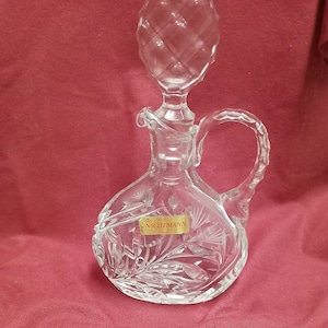 May include: A clear glass decanter with a stopper and a handle. The decanter has a floral etched design and a label that reads "Nachtmann."