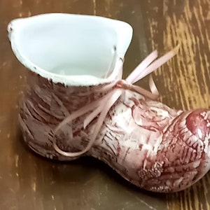 May include: A small, pink and white ceramic boot with a pink ribbon tied around it. The boot has a distressed look and is designed to hold small items.