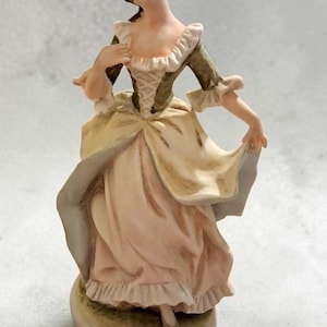 May include: A porcelain figurine of a person wearing a period-style dress with a dark green bodice, a pale yellow overskirt, and a light pink underskirt. The figure has brown hair, ruffled sleeves, and stands on a circular base with a light finish.