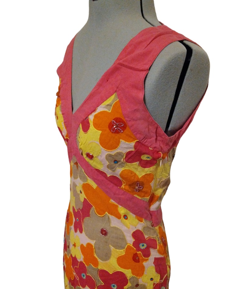 Puede incluir: Vestido sin mangas con escote en V, con un vibrante estampado floral. El vestido presenta una combinaci&oacute;n de flores amarillas, naranjas, rosas y marrones, con detalles de cuentas. El corpi&ntilde;o y las correas son de color rosa s&oacute;lido, creando un contraste llamativo.