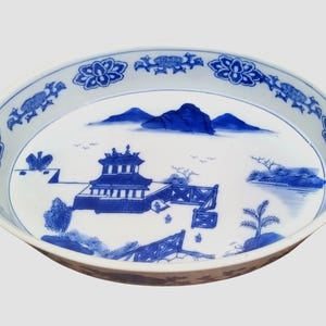 May include: A white ceramic plate with a blue and white design of a Chinese landscape with a pagoda, mountains, trees, and a river. The plate has a scalloped edge and a blue and white floral border.