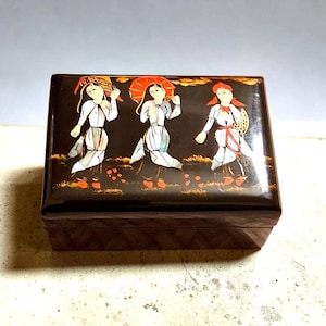 May include: A rectangular black lacquer box featuring a lid decorated with an inlaid scene of three figures in white garments, one holding a red umbrella, set against a dark background with orange accents and gold-toned highlights.