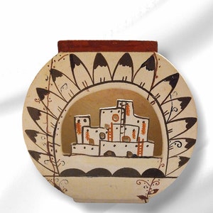 May include: A ceramic vase with a brown rim and a white background. The vase is decorated with a black and white design of feathers and a pueblo-style building in the center.