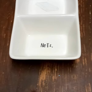 Puede incluir: Plato de cerámica blanco con dos compartimentos. El compartimento superior tiene un dibujo de un trozo de papel. El compartimento inferior dice "Note."