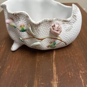 May include: A white ceramic bowl with a scalloped rim and three feet. The bowl is decorated with pink and green flowers and gold accents.