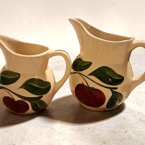 Puede incluir: Dos jarras de crema de cerámica con un esmalte beige. Cada jarra presenta un diseño de cerezas con cerezas rojas y hojas verdes. Las jarras tienen un asa curva y un pico para verter.