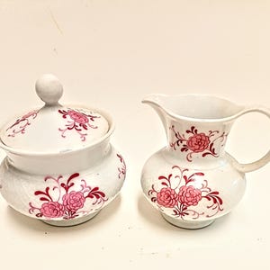 May include: A white ceramic sugar bowl with a lid and a small creamer pitcher. Both pieces are decorated with pink floral designs and dark red accents. The sugar bowl has a textured surface, and the creamer has a handle and spout.