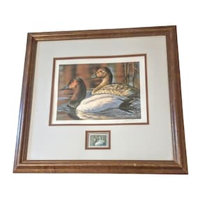 May include: Framed print featuring two ducks on water, with a smaller print of a duck below. The ducks have brown and white feathers, set against a backdrop of reeds and water. The frame is a warm brown color.