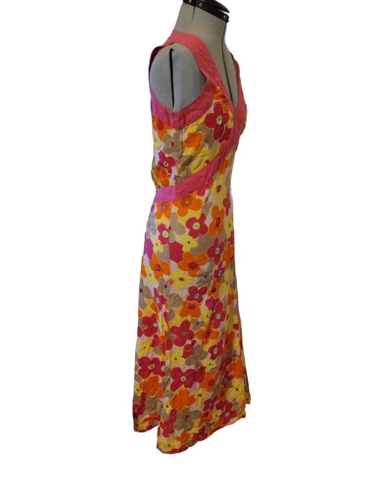 Puede incluir: Un vestido largo sin mangas con un estampado floral en tonos amarillos, naranjas, rosas y marrones. El vestido tiene un escote en V y ribetes rosas. El vestido se exhibe en un maniqu&iacute;.
