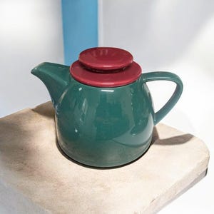 May include: A ceramic teapot with a dark green body and a burgundy lid. The teapot has a rounded shape with a curved handle and a spout. The lid is composed of two stacked, circular pieces. The teapot is sitting on a light-colored, textured surface.