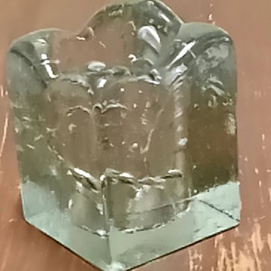 Puede incluir: Un portavelas de vidrio transparente con base cuadrada y superficie texturizada. El portavelas tiene una parte superior única, ligeramente curvada.