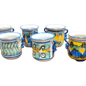 Puede incluir: Seis pequeñas tazas de cerámica con asas azules y diseños florales coloridos. Las tazas son blancas con detalles azules, amarillos y verdes.