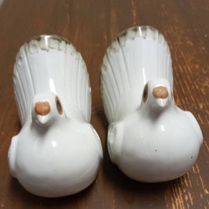 Puede incluir: Dos figuritas de palomas blancas de cerámica con ojos y picos marrones. Las palomas tienen un diseño moteado marrón y blanco en sus colas.