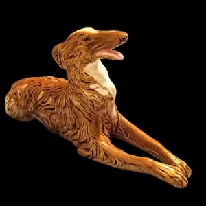 May include: A ceramic sculpture of a dog in a reclining pose. The dog is primarily brown with a white chest and a pink tongue. The sculpture has detailed fur texture and is set against a black background.