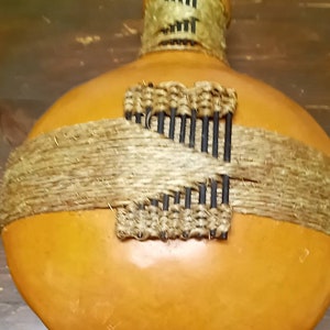 May include: A gourd bottle with a woven design and a brown, natural fibre band around the middle. The bottle is a light orange colour.