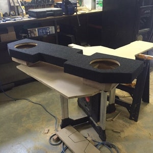 Może przedstawiać: Czarna obudowa subwoofera samochodowego z dwoma otworami na głośniki. Obudowa stoi na drewnianym stole w warsztacie.