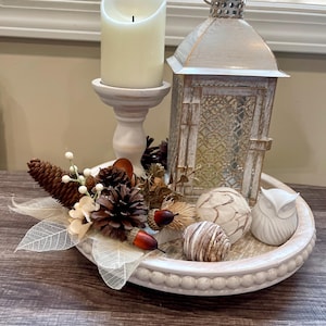 May include: A round beaded tray holding a white pillar candle on a pedestal, a metal lantern with a distressed finish, a small white owl figurine, pinecones, artificial acorns, and decorative skeleton leaves arranged as a centerpiece.