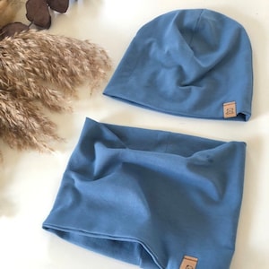 Könnte beinhalten: Ein blaues Mütze- und Halstuch-Set. Beide Artikel sind aus weichem Stoff gefertigt und haben ein kleines, quadratisches Etikett mit einer Bärenillustration. Das Set wird auf einer weißen Oberfläche mit getrockneten Pflanzen im Hintergrund präsentiert.