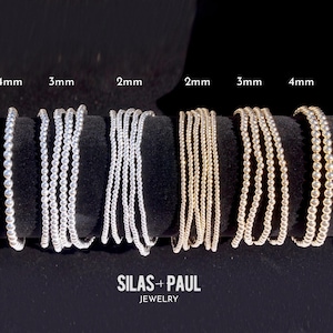 Könnte beinhalten: Präsentation von silbernen und goldenen Perlenarmbändern in den Größen 2 mm, 3 mm und 4 mm. Die Armbänder sind auf einer schwarzen Oberfläche angeordnet, mit dem Markennamen "SILAS + PAUL JEWELRY".