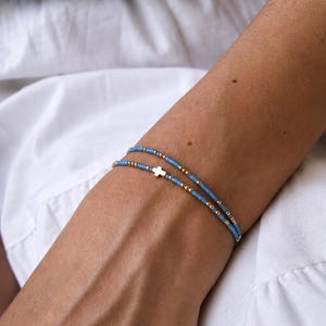 May include: A double-strand bracelet featuring small light blue disc beads interspersed with tiny metallic gold-tone beads. One strand includes a small, centered metallic gold-tone cross charm. The bracelet is worn on an arm against a white fabric.